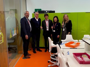 A group of five people standing in a brightly colored science lab with orange flooring and green cabinets, holding laboratory pipettes. They wear ID badges and are dressed in professional attire, indicating a formal visit. The lab features trays of scientific equipment, safety goggles, and illustrated glassware on the wall, suggesting a STEM-focused educational setting.