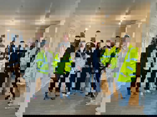 A group of people, including construction staff in high‑visibility jackets and others in everyday clothing, stand together inside a newly completed, unfurnished home with light wooden flooring and white walls.