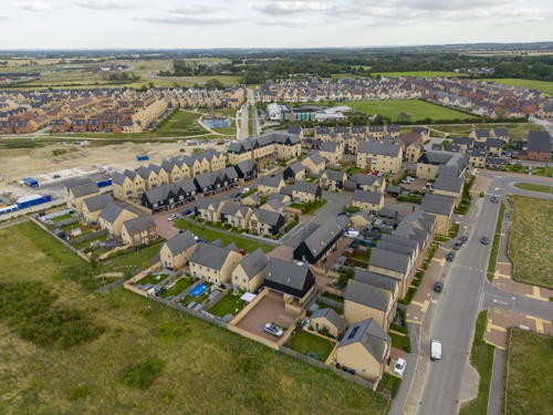 An aerial shot of the new town of Northstowe. There are lots of homes surrounded by green fields, under a cloudy sky.