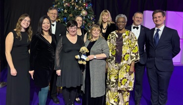 A group of people dressed in formal attire standing on a purple carpet in front of a decorated Christmas tree with blue and gold ornaments. The setting features festive decorations, including golden star patterns and string lights, creating a celebratory atmosphere. One person holds a small stuffed dog toy.