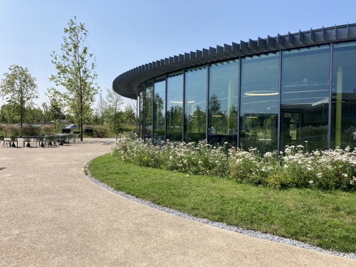 An image showing Melbourn Science Park. There is a round glass-fronted building surrounded by planted shrubs and some trees.